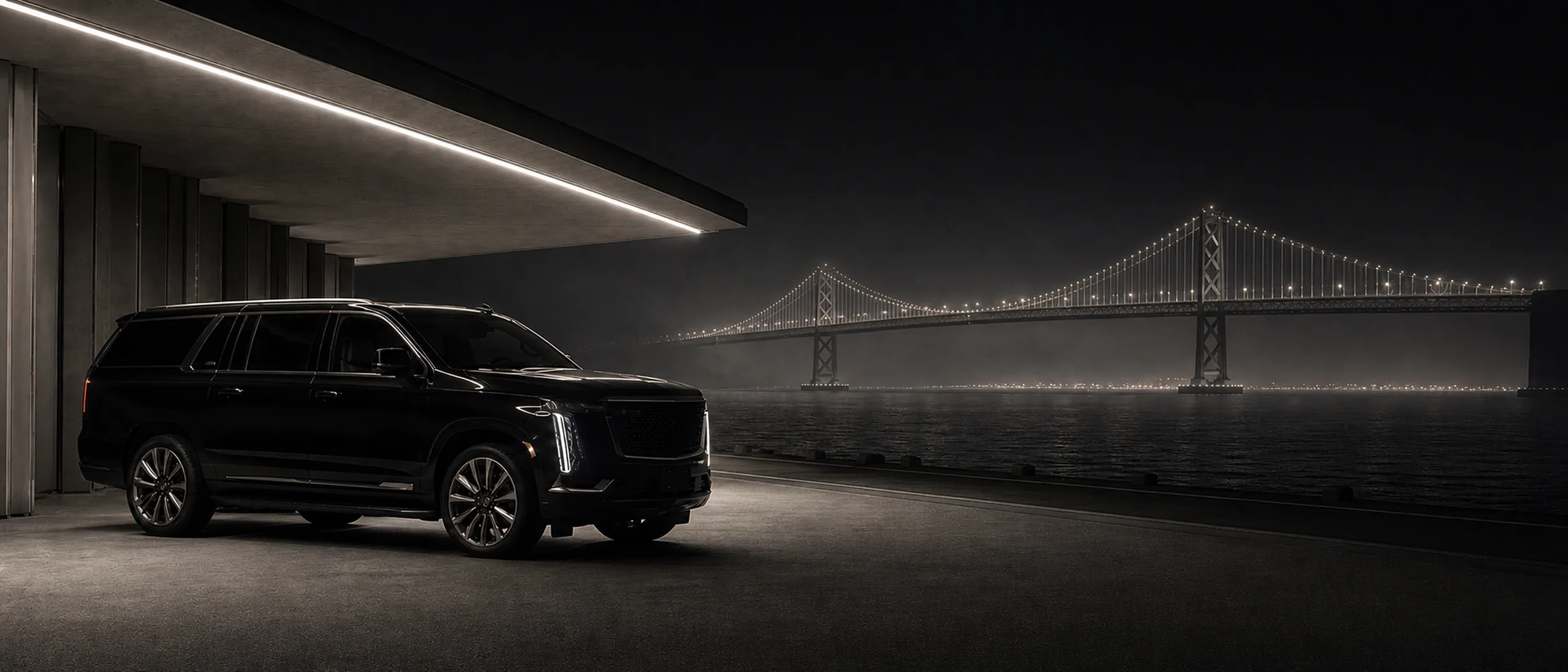 A black Cadillac Escalade parked under a covered driveway with the Bay Bridge glowing across the water at night.